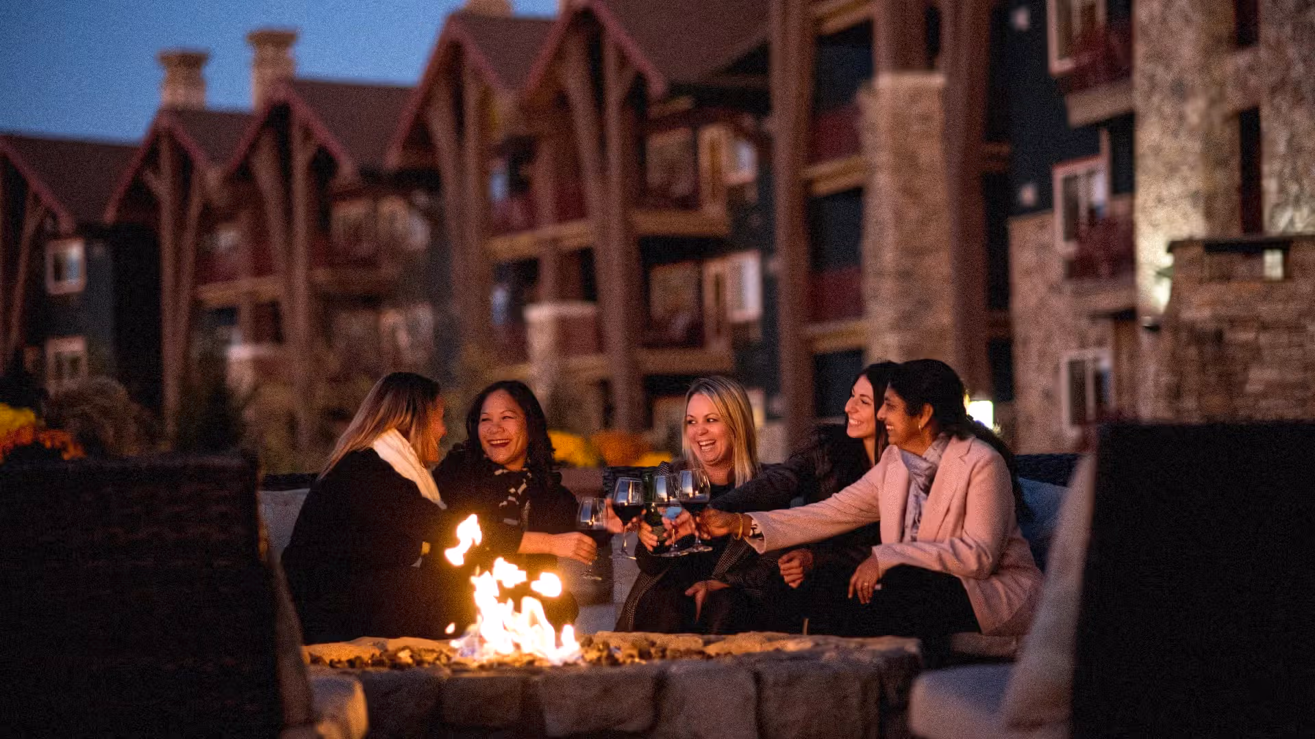 Girlfriends sit around firepit on Fire & Water Terrace and cheers with wine.