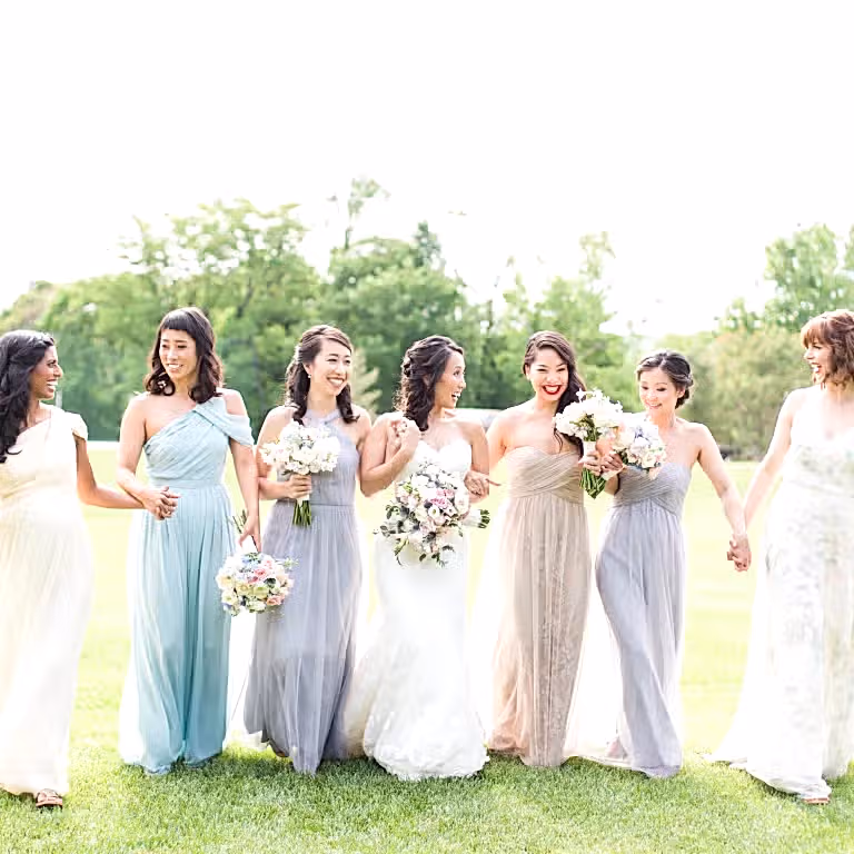 Bridesmaids and bride walk through grass field.